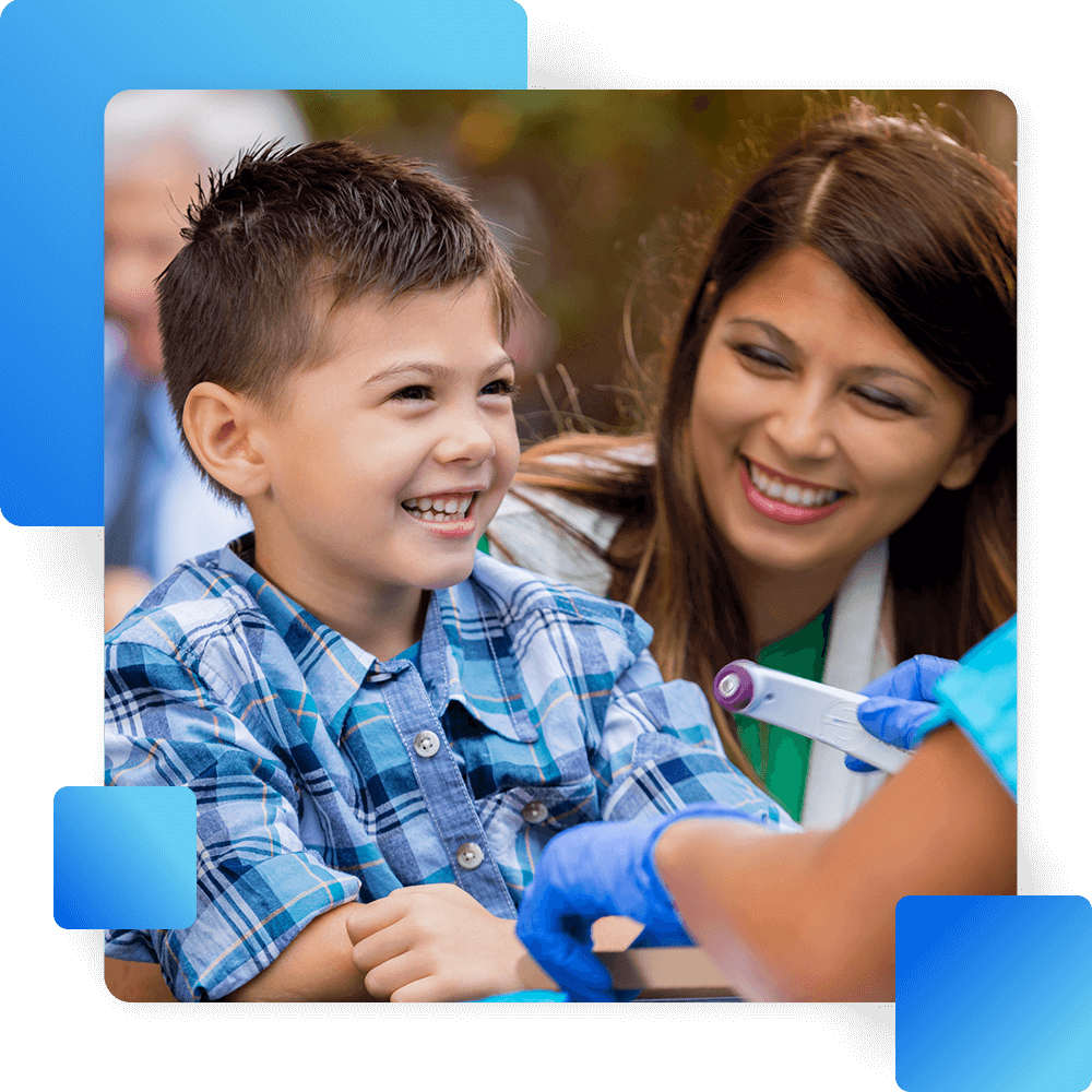 A smiling boy in a plaid shirt receives a medical checkup from a healthcare worker, while a woman smiles in the background.